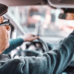 Senior man with hat and eyeglasses adjusting rear mirror while sitting in his car. Other hand on steering wheel. Picture taken from back seat.