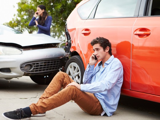 young man sitting next to car, talking on the phone after a car accident