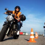 Female student with helmet taking motorcycle lessons and practicing ride. In background traffic cones and instructor with checklist rating and evaluating the ride. Motorcycle school of driving.